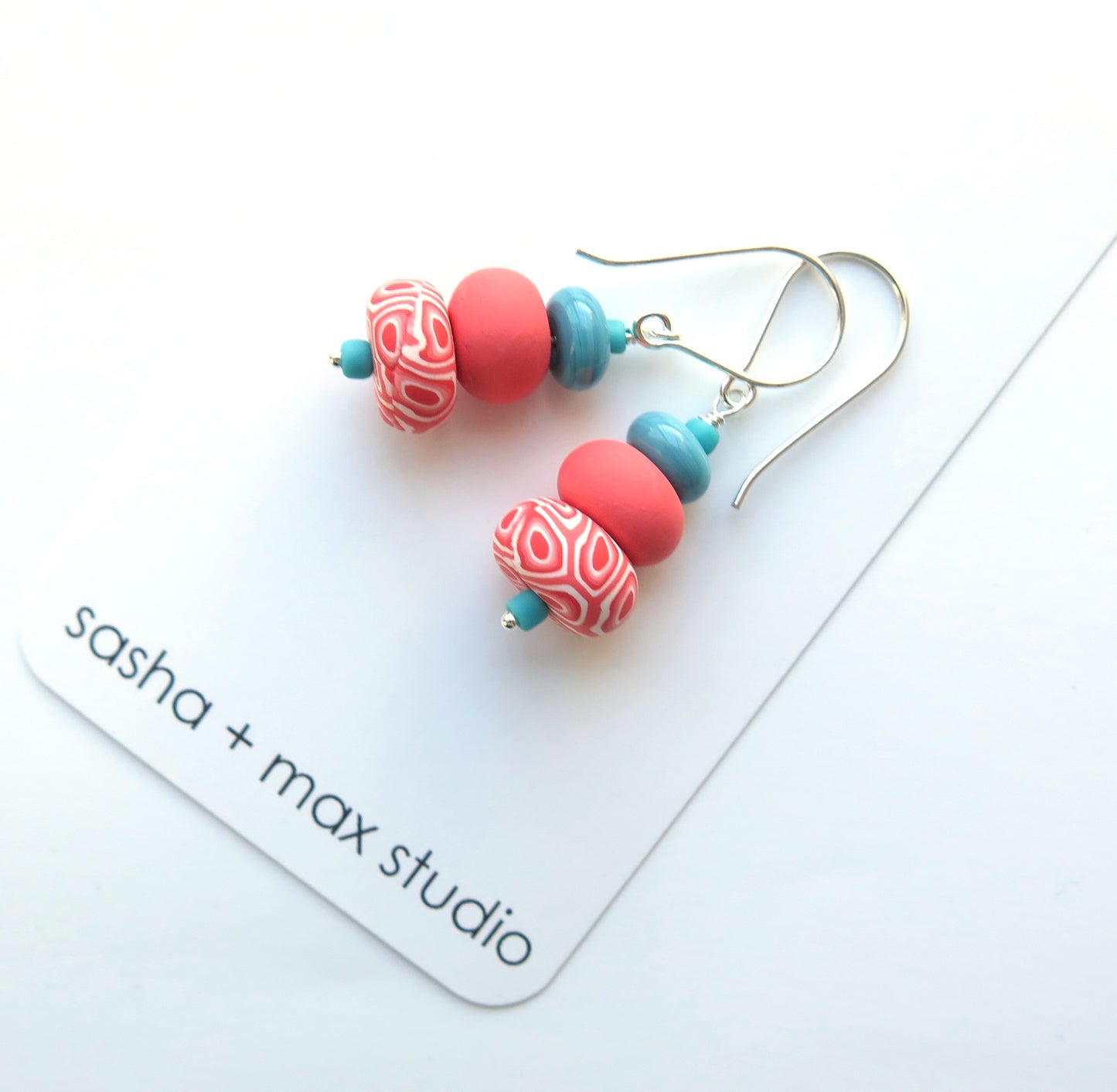 Earrings with red and blue beads on a white card with 'sasha + max studio' branding.